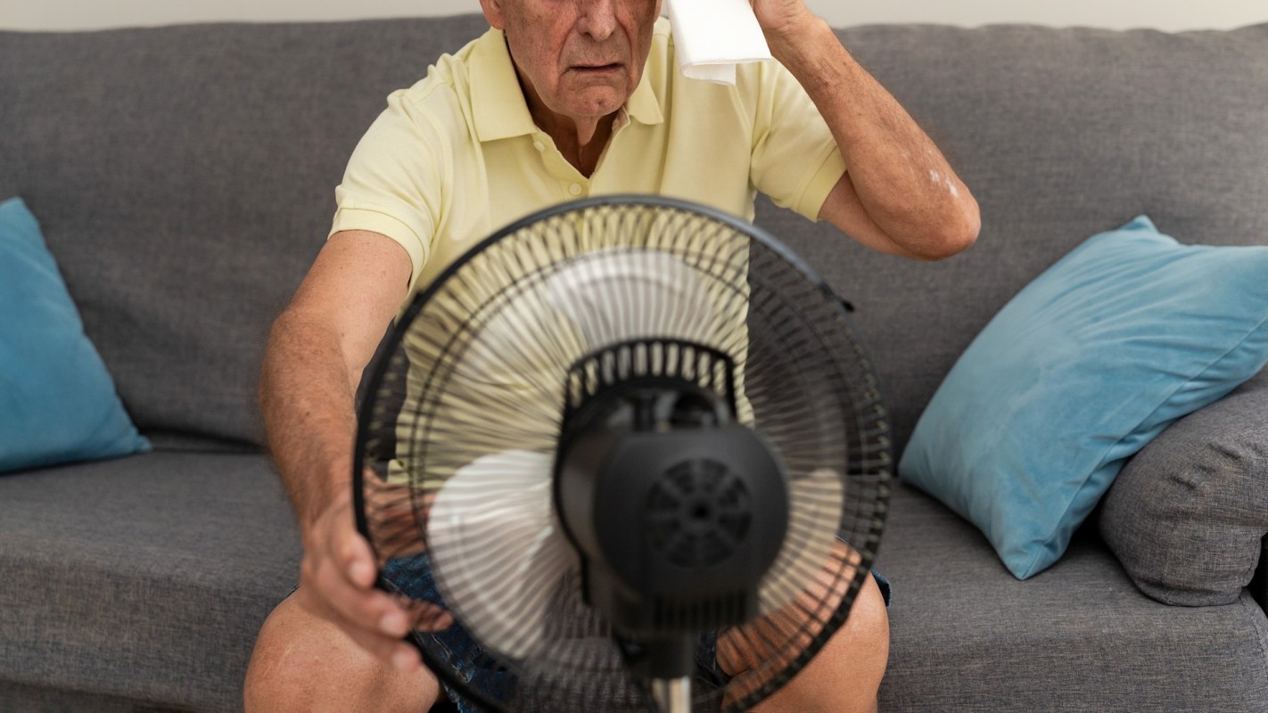 An older man sits on a couch, wiping his forehead with a towel while a fan blows air toward him.