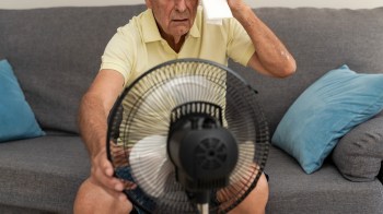 An older man sits on a couch, wiping his forehead with a towel while a fan blows air toward him.
