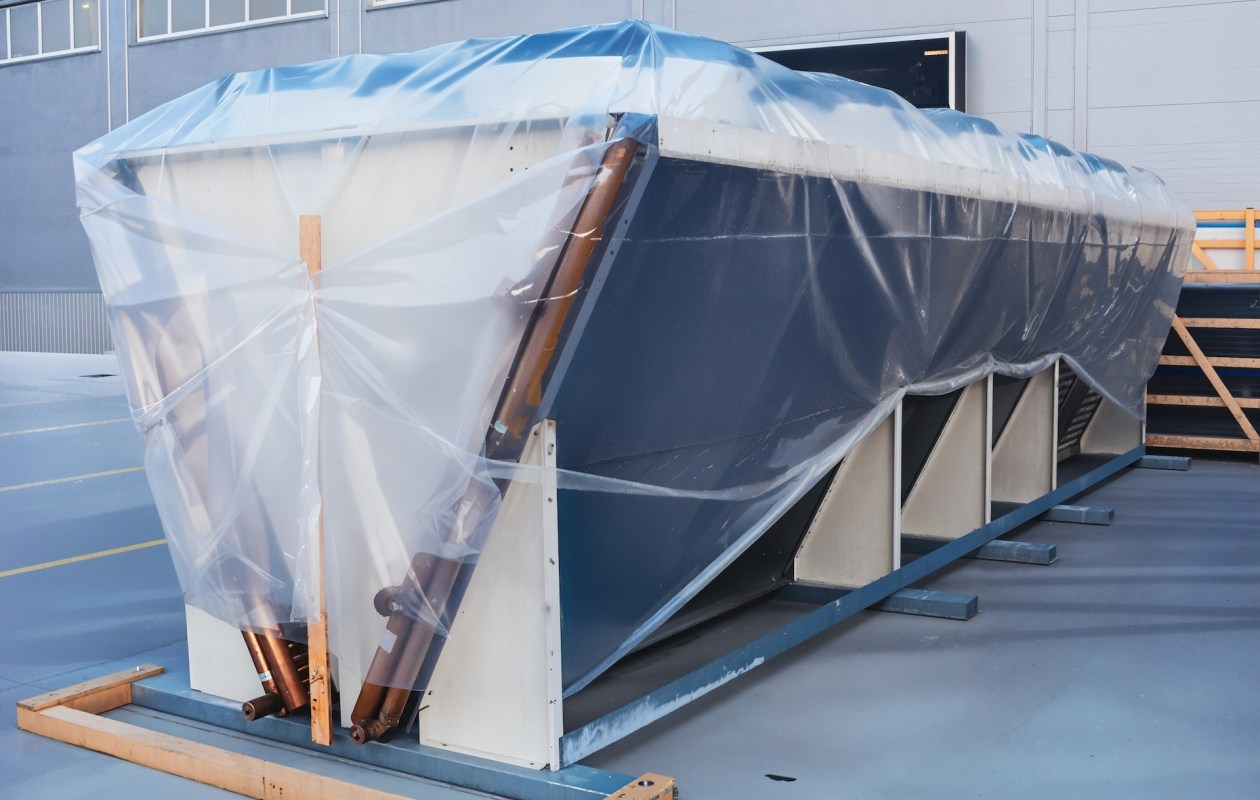 A large object covered with clear plastic sheeting, secured on a wooden frame in an industrial setting.