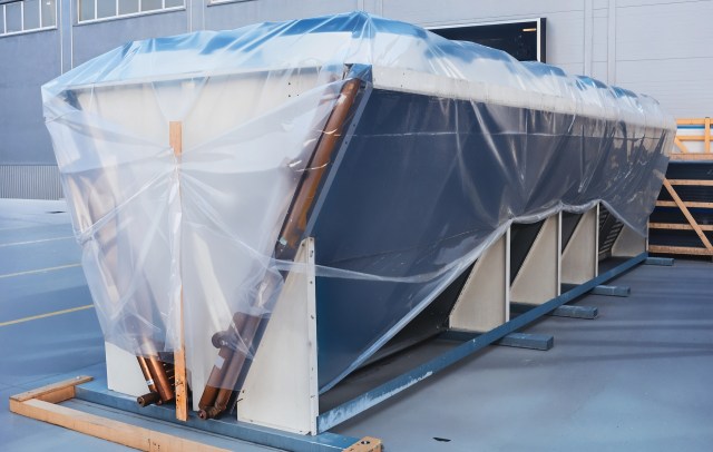A large object covered with clear plastic sheeting, secured on a wooden frame in an industrial setting.