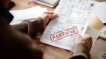 A person holds a bill marked "PAST DUE" on a wooden table surrounded by other documents.