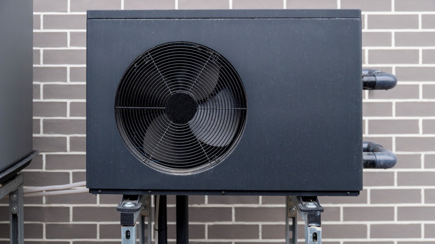 An outdoor air conditioning unit mounted on metal supports against a brick wall.