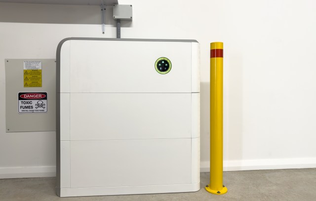 A large white storage unit next to a yellow safety bollard and warning signs on the wall.