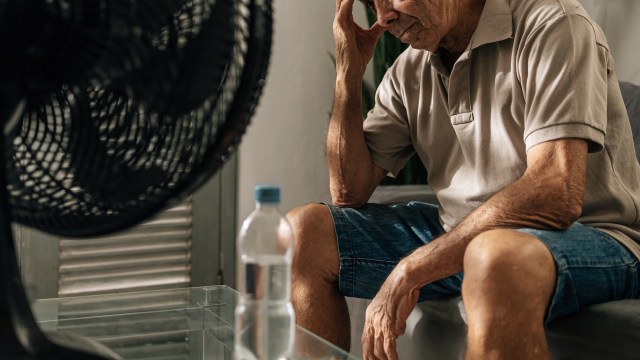 An elderly man sitting next to a fan, with a bottle of water on a table in front of him.