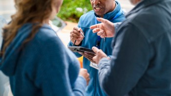 A salesman at the door discussing something with a tablet and a pen in hand to two people.