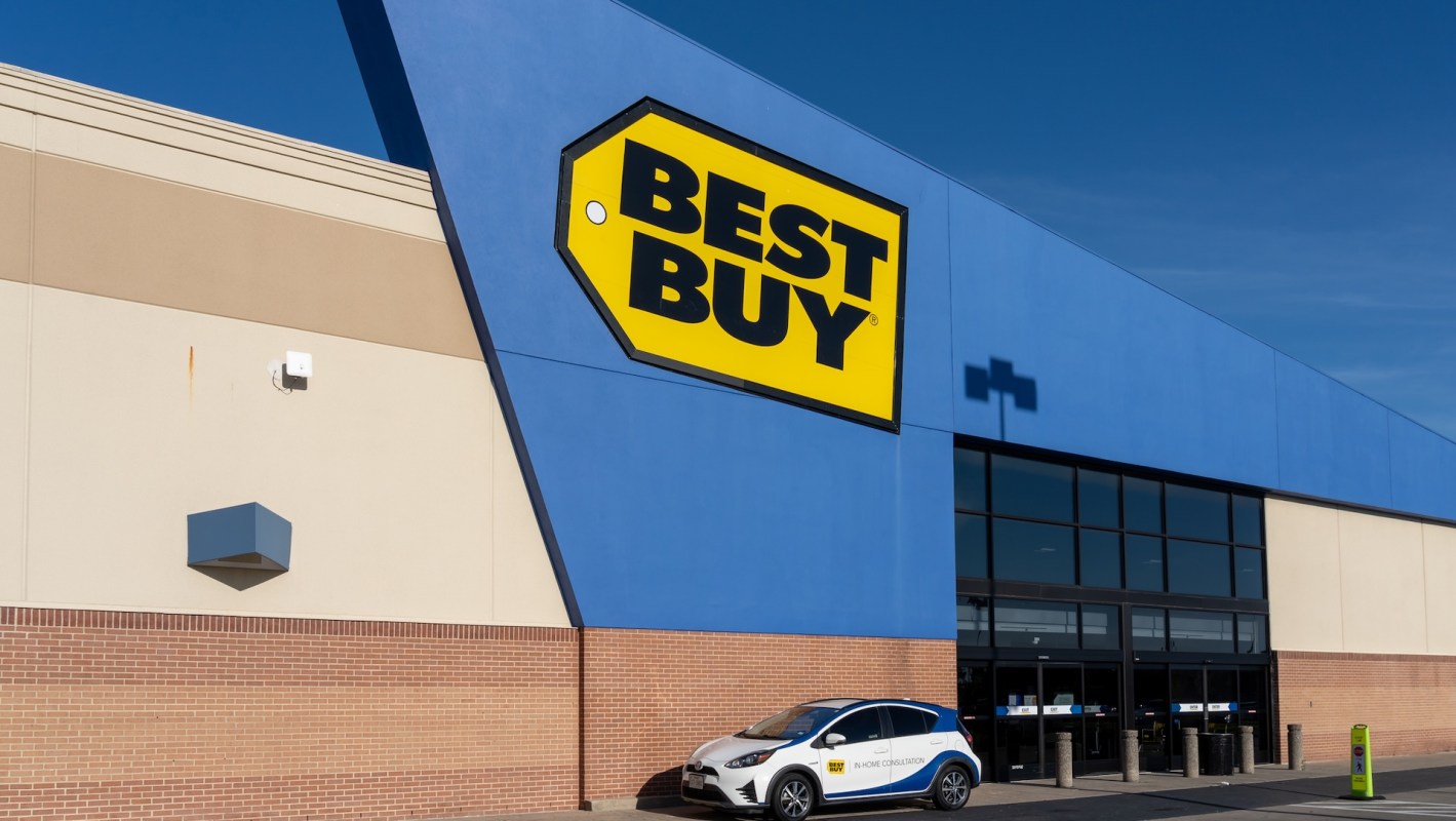 The exterior of a Best Buy store with a blue wall and a parked vehicle displaying an in-home consultation advertisement.