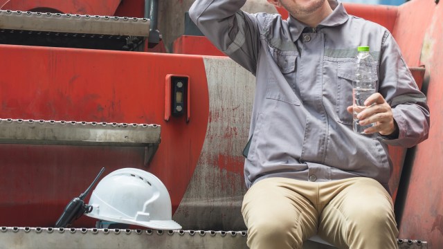 A worker in a gray uniform sits on steps, holding a water bottle next to a white hard hat and a radio.
