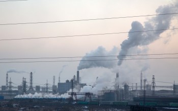 An industrial landscape with buildings and smokestacks emitting white smoke against a gray sky.