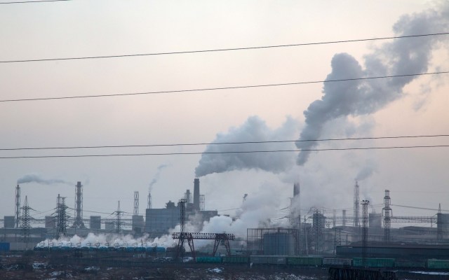 An industrial landscape with buildings and smokestacks emitting white smoke against a gray sky.
