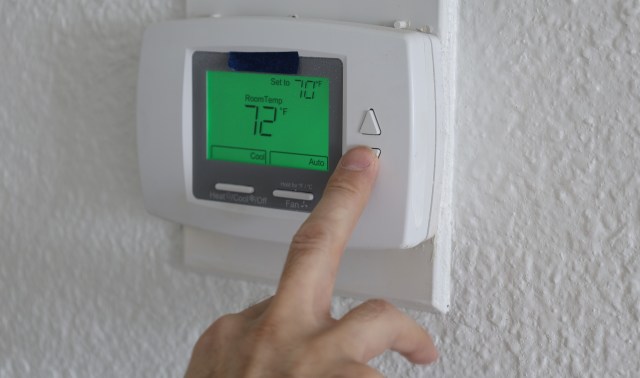 A person adjusting a digital thermostat displaying the current and set room temperatures.