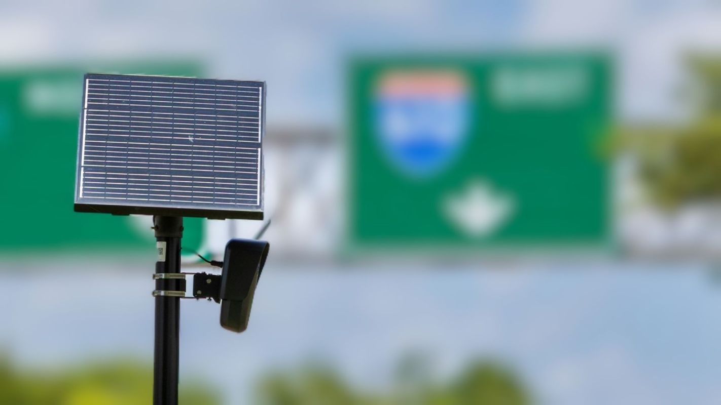 A solar panel mounted on a pole with green highway signs blurred in the background.
