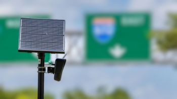 A solar panel mounted on a pole with green highway signs blurred in the background.
