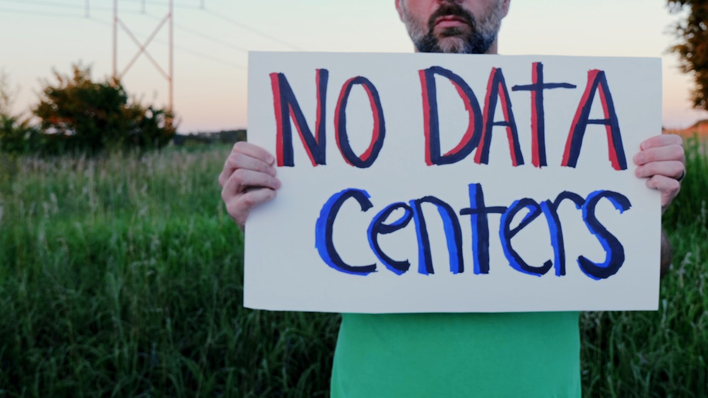 A person holds a sign that reads "NO DATA CENTERS" in a field at sunset.