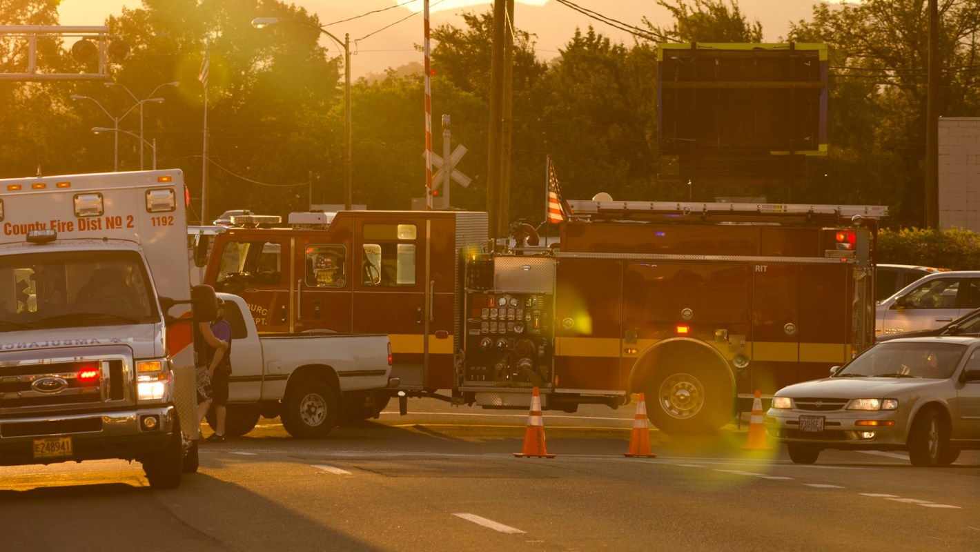 Emergency vehicles, including an ambulance and fire truck, are blocking traffic at a railroad crossing during sunset.