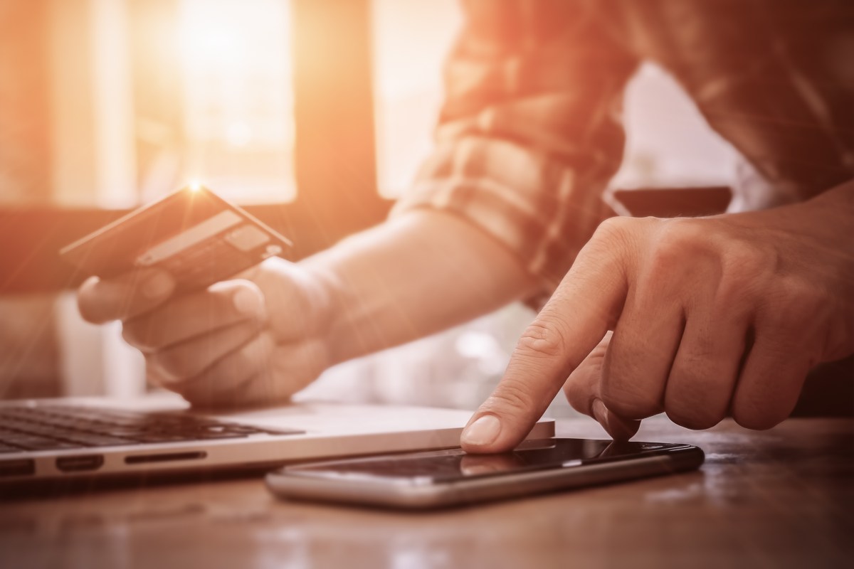 A person holds a credit card while using a laptop and a smartphone on a table.
