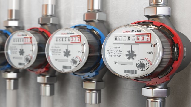 Three water meters are mounted on a wall, showing various readings and connected by metal pipes.
