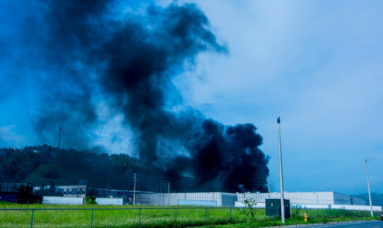 A plume of black smoke rises from a building near a grassy area.