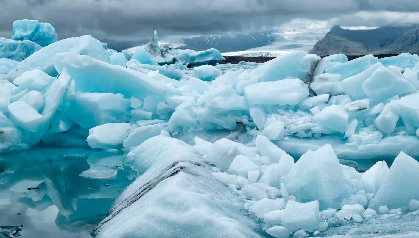 An ice-covered landscape with floating ice chunks and glacial water under a cloudy sky.
