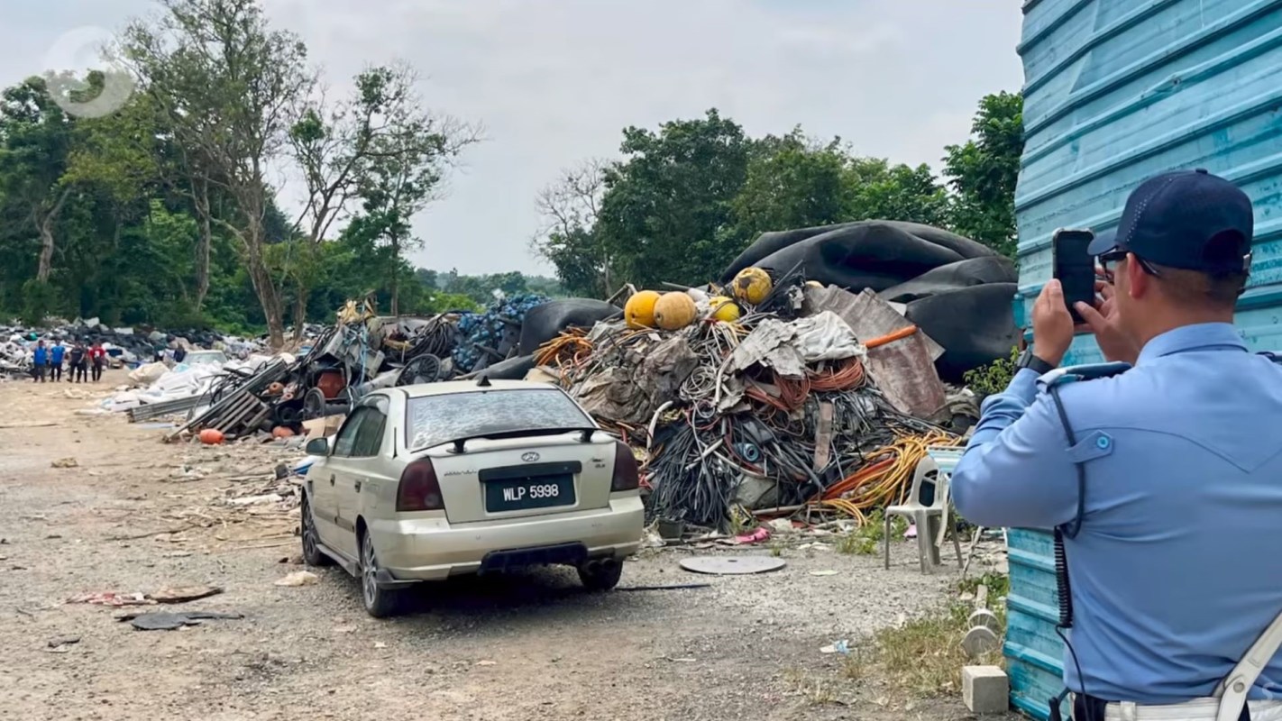 A man takes a photo near a large pile of discarded materials and an abandoned car.