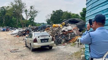 A man takes a photo near a large pile of discarded materials and an abandoned car.