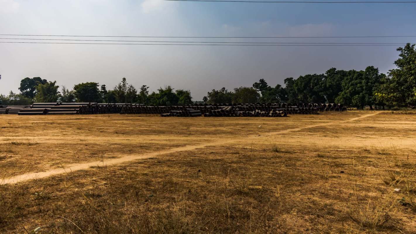 A dry field with stacked pipes and scattered trees in the background under a cloudy sky.