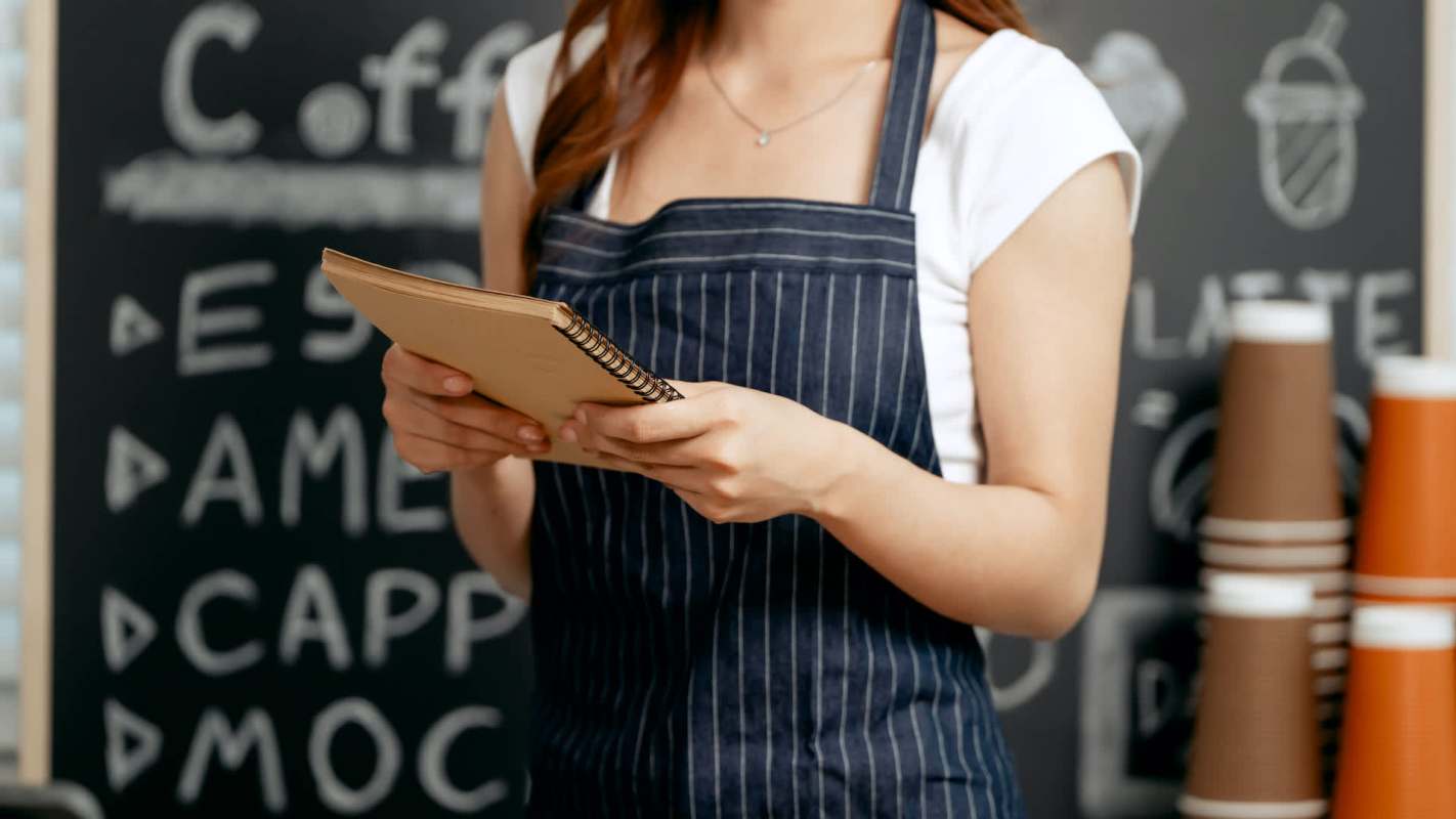 A person in an apron holds a notebook in front of a chalkboard menu.