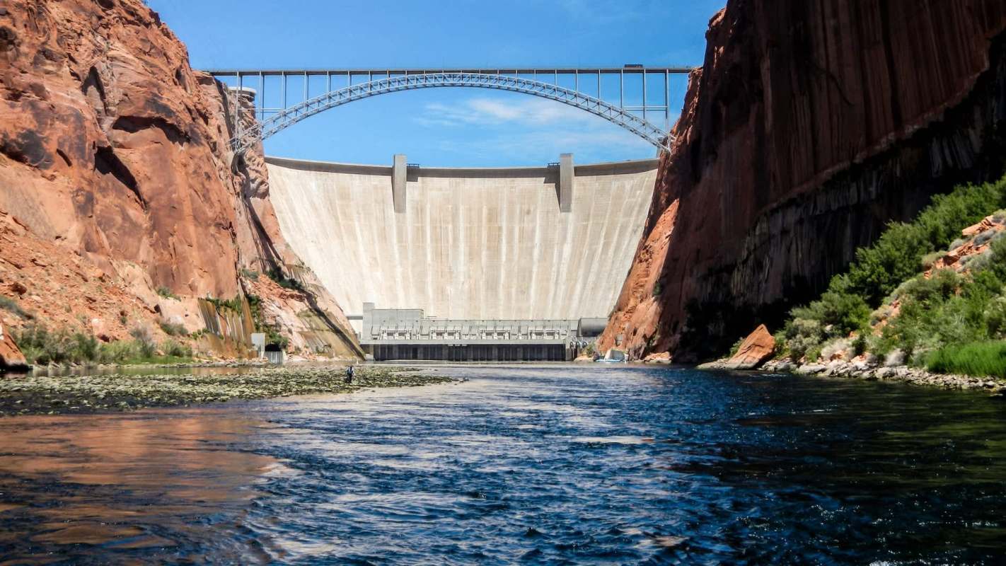 Glen Canyon Dam surrounded by rocky cliffs, with a river in the foreground.