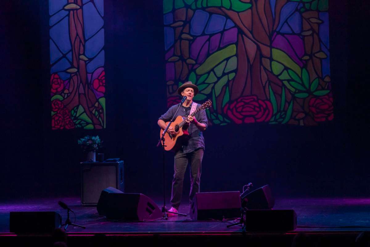 A musician plays guitar on stage in front of colorful stained glass backdrops.