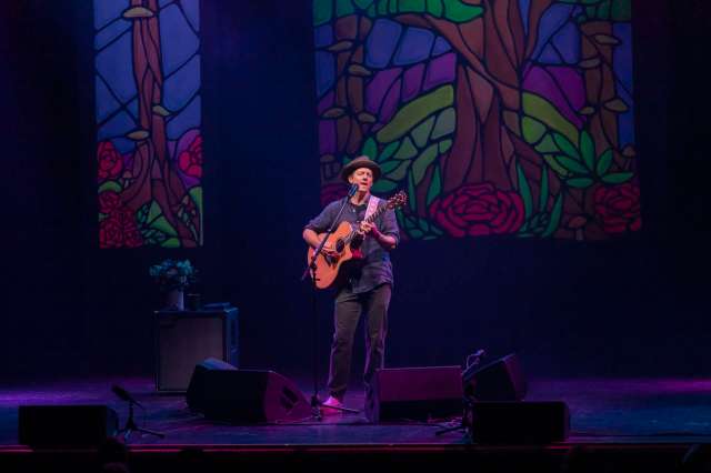 A musician plays guitar on stage in front of colorful stained glass backdrops.