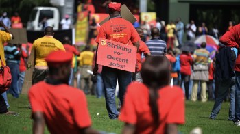 A crowd of protesters wearing red shirts gathers, some holding signs for better job conditions.
