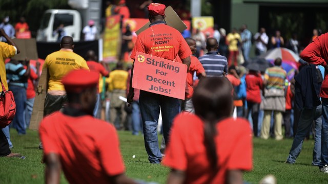 A crowd of protesters wearing red shirts gathers, some holding signs for better job conditions.