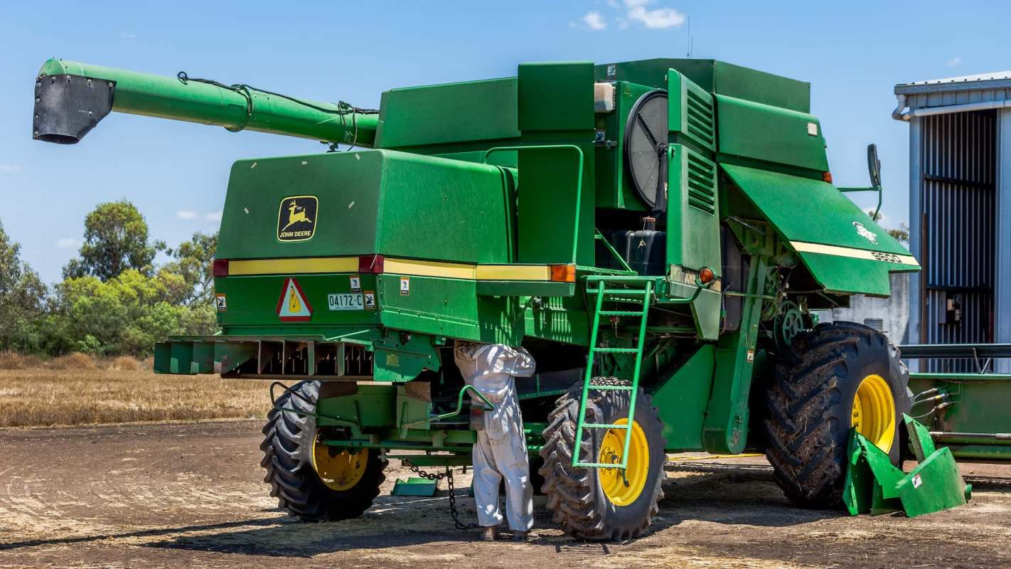 A green John Deere harvesting machine with a worker inspecting it in a farm setting.