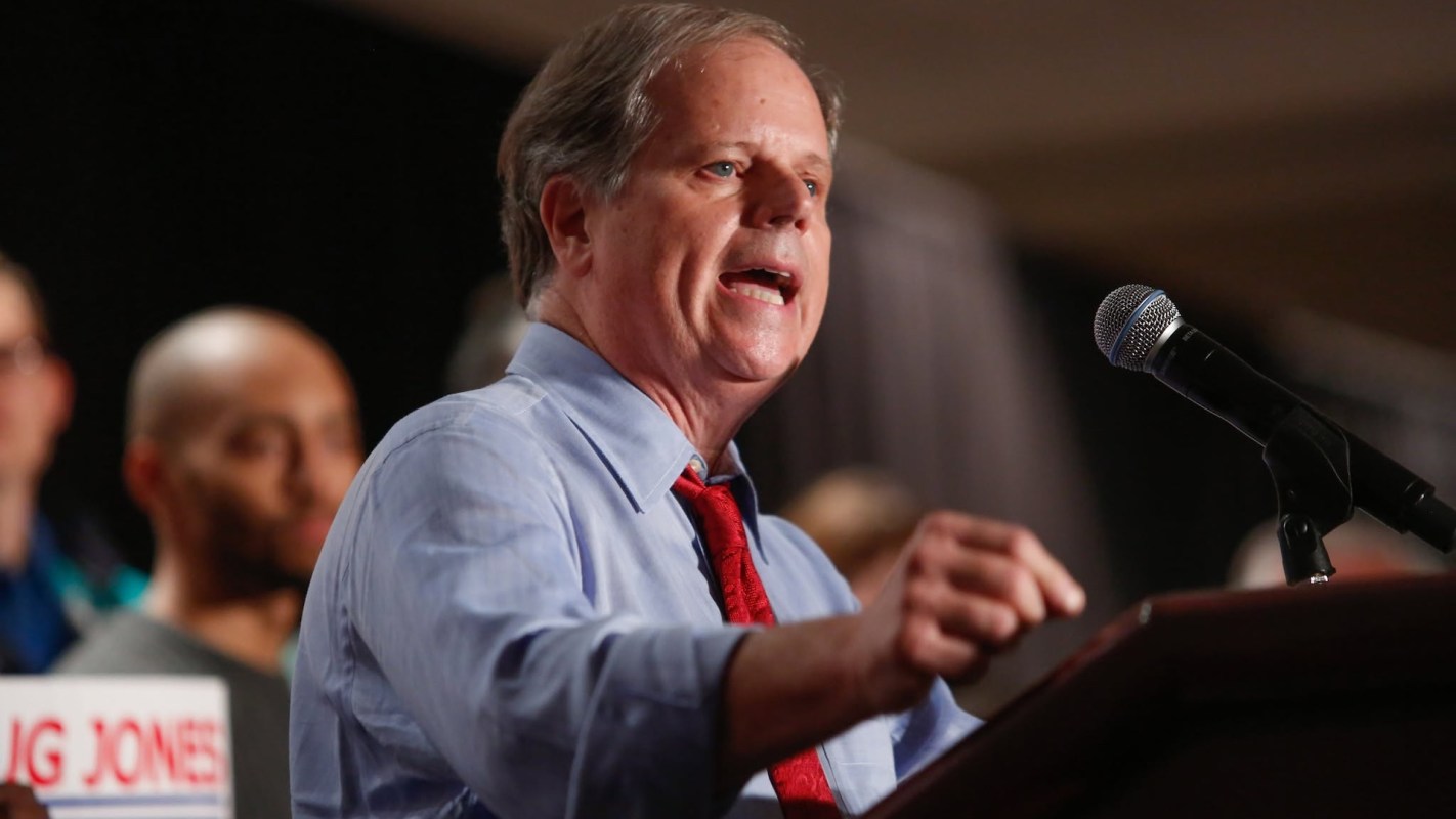 Doug Jones speaks at a podium with a microphone, with supporters visible in the background.