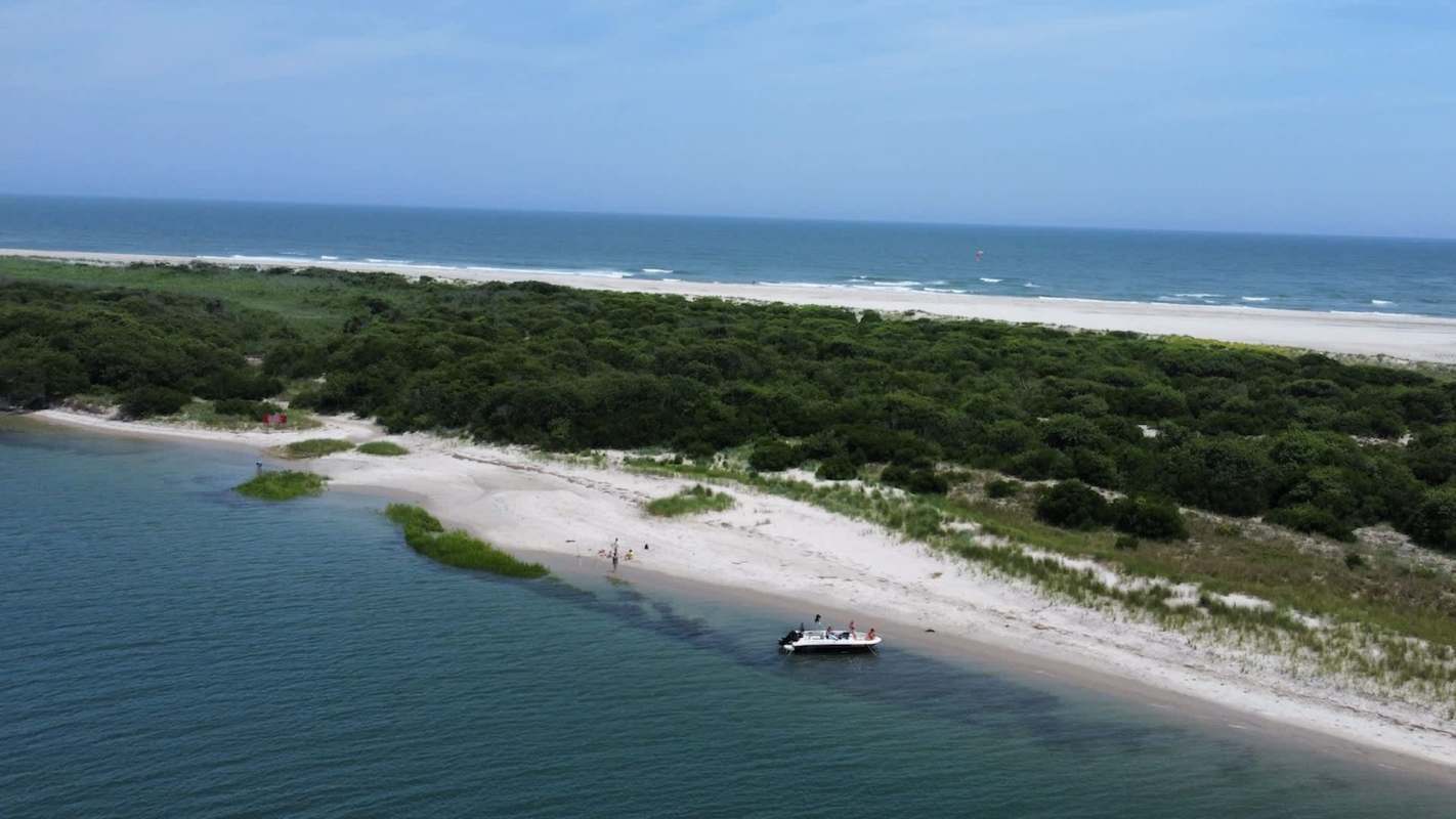 A boat is anchored near a sandy beach with green vegetation and the ocean in the background.