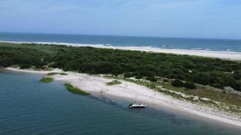 A boat is anchored near a sandy beach with green vegetation and the ocean in the background.