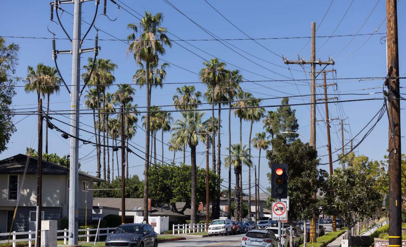 A street lined with palm trees and utility poles, with traffic lights and parked cars visible.