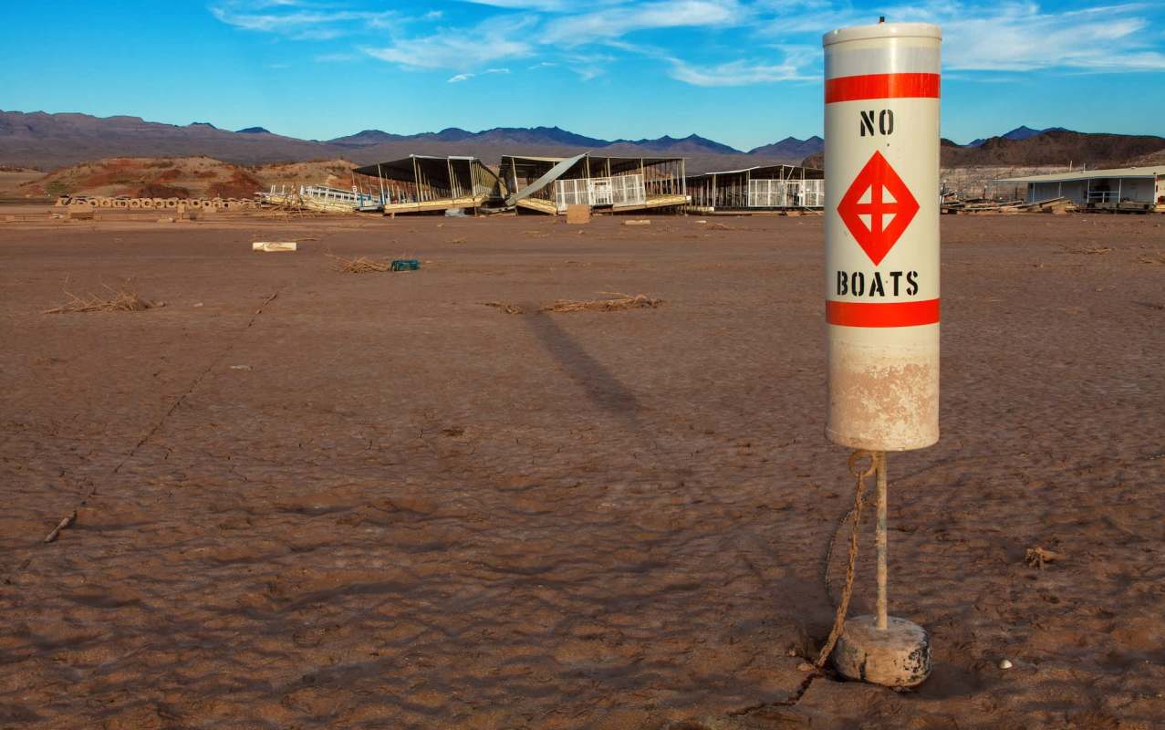 A "No Boats" sign stands in a dry, cracked Lake Mead with abandoned structures in the background.