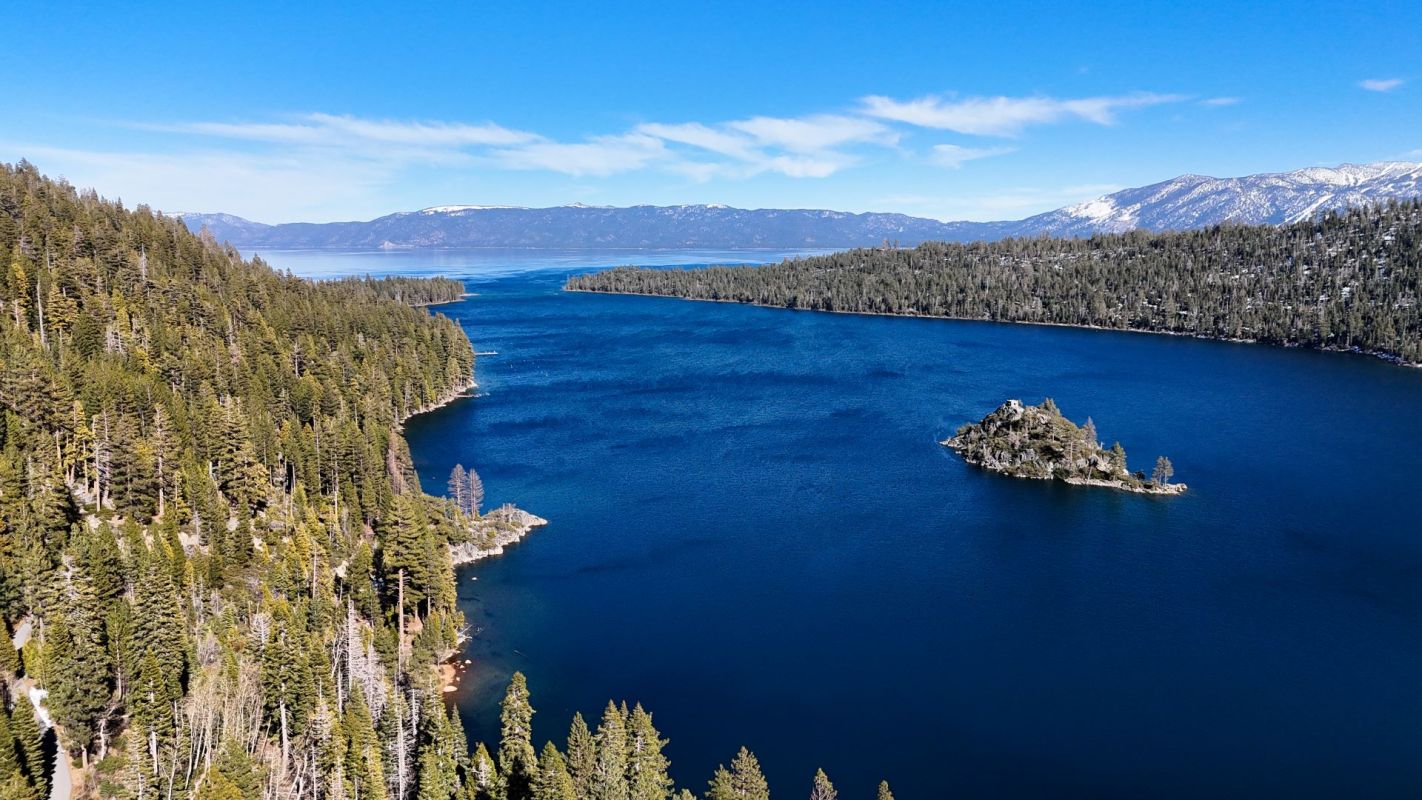 A panoramic view of a lake surrounded by forested mountains under a clear blue sky.
