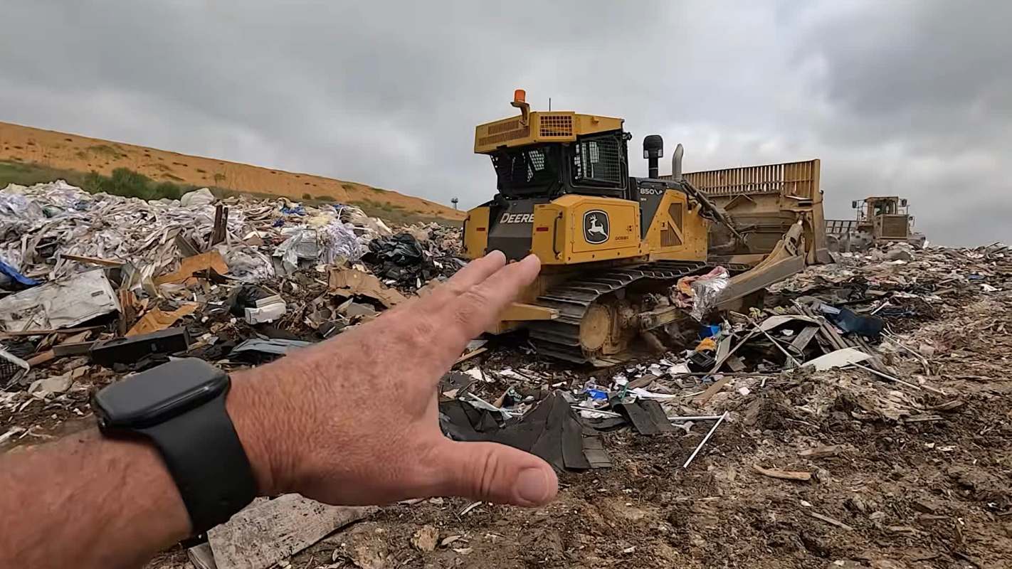 A hand gestures towards a bulldozer working at a landfill covered in piles of debris and waste.