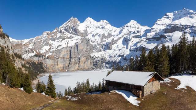 A snowy mountain landscape with a lake and a wooden cabin surrounded by evergreen trees.