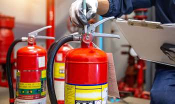 A person inspects a fire extinguisher while holding a clipboard in a maintenance area.