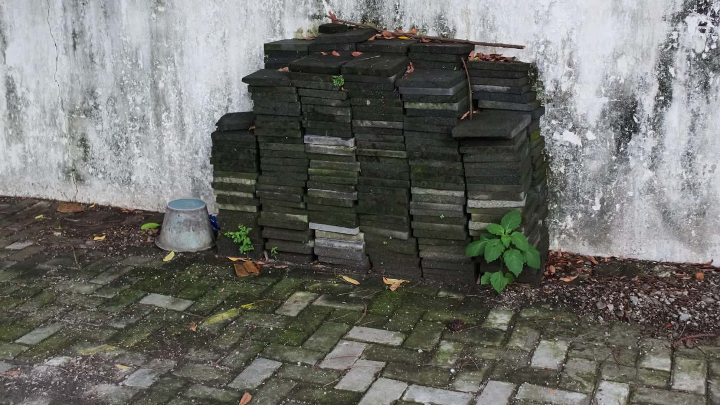 A stack of gray paving stones next to a concrete wall with green plants and a metal bucket on the ground.