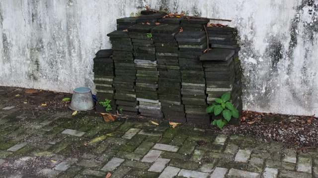 A stack of gray paving stones next to a concrete wall with green plants and a metal bucket on the ground.