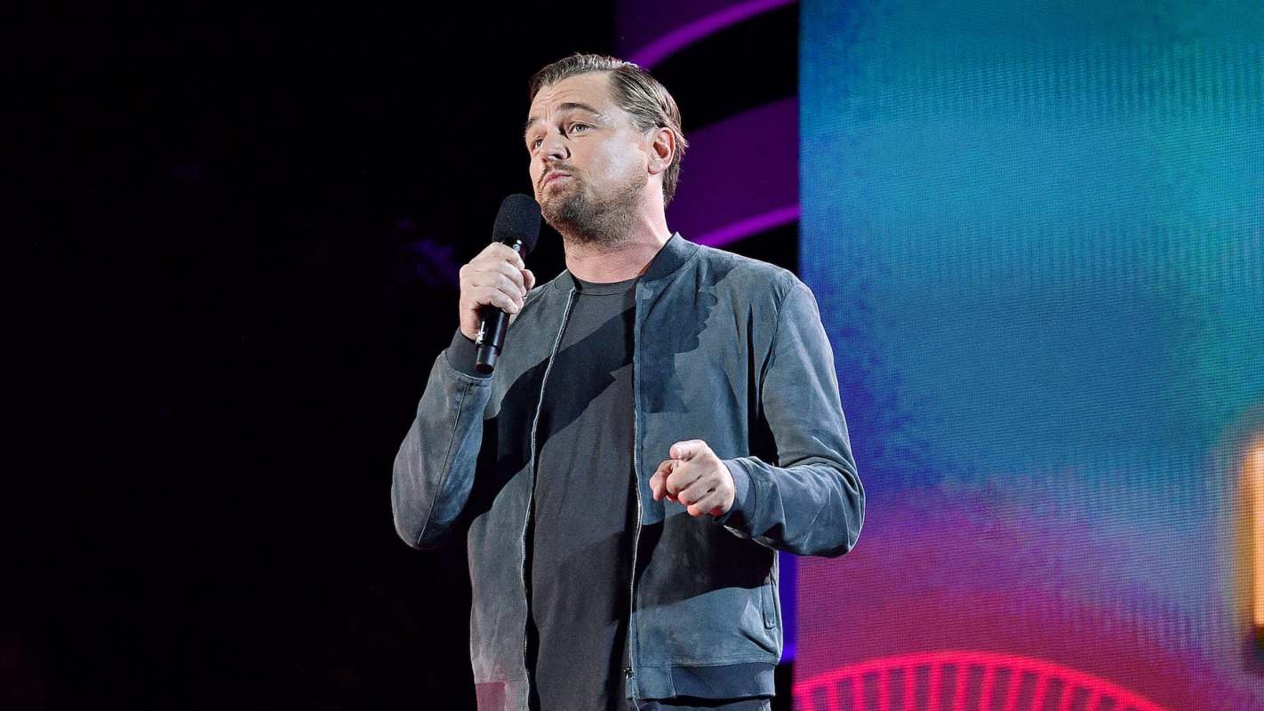 Leonardo DiCaprio speaks into a microphone on stage with a colorful background.