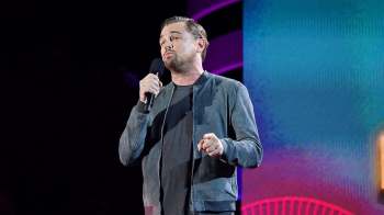 Leonardo DiCaprio speaks into a microphone on stage with a colorful background.