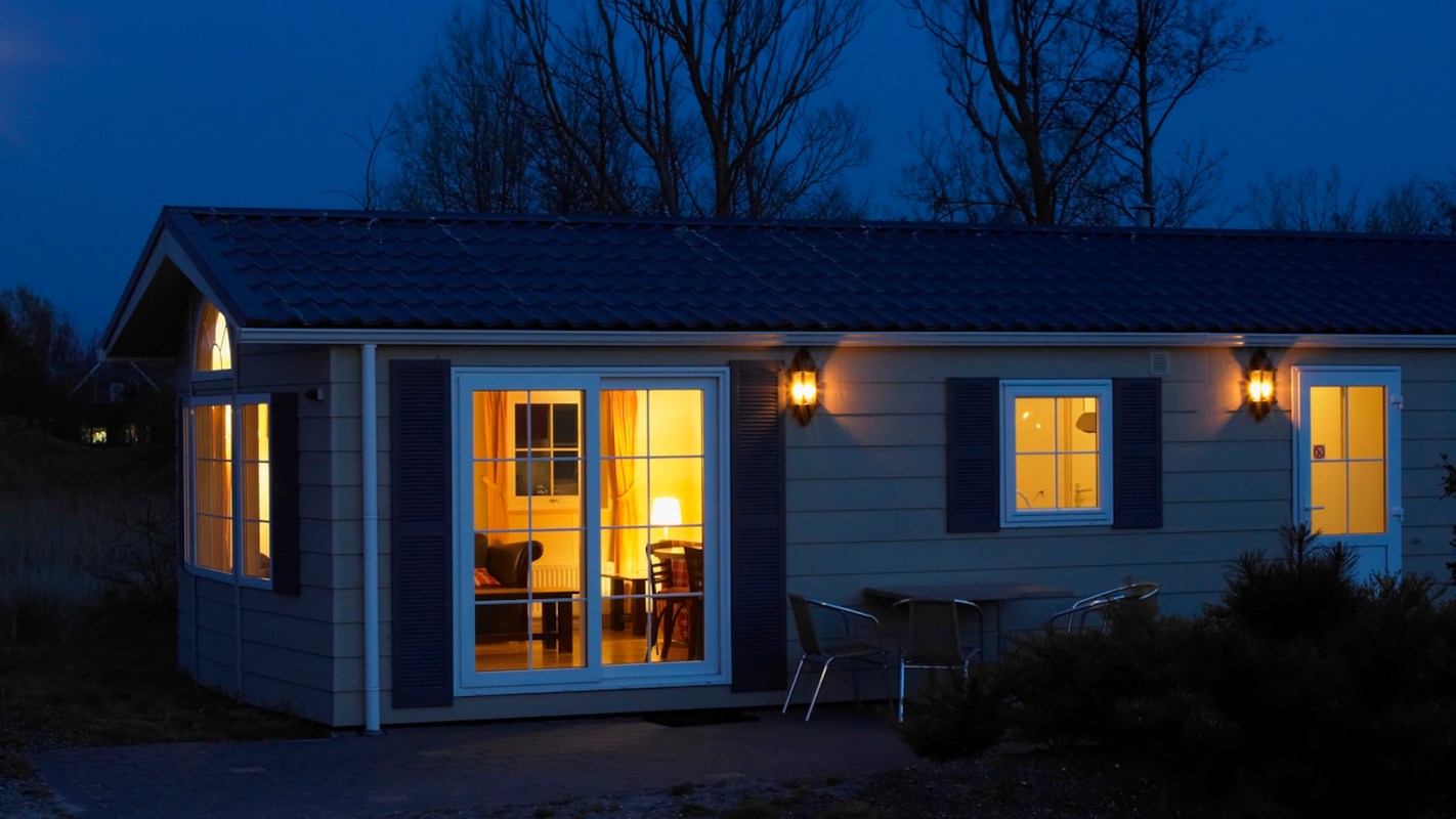 A lit cabin at night with visible furniture through the windows and outdoor seating.