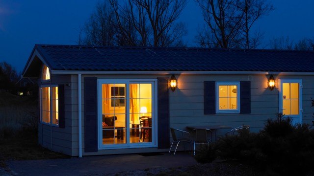 A lit cabin at night with visible furniture through the windows and outdoor seating.