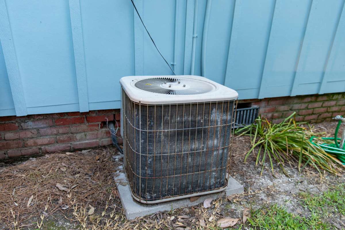 An air conditioning unit next to a blue wall and brick foundation, surrounded by dry grass and plants.