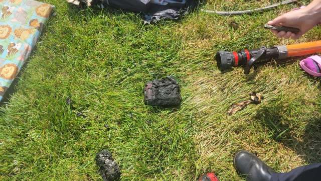 A blackened object is on the grass next to a fire hose and various items, including a colorful blanket and footwear.