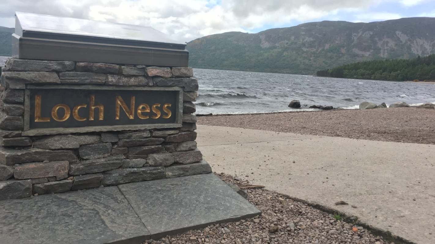 A stone sign reading "Loch Ness" by the lakeshore with waves and hills in the background.